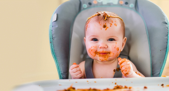 Lächelndes Baby mit Essensresten im Gesicht und auf dem Kopf im Hochstuhl.
