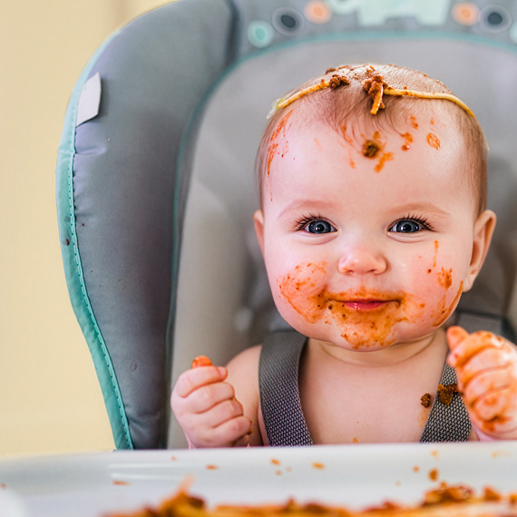Lächelndes Baby mit Essensresten im Gesicht und auf dem Kopf im Hochstuhl.