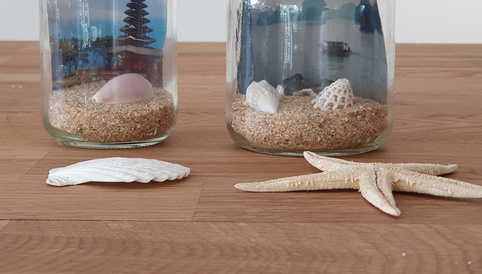 Zwei dekorative Gläser mit Muscheln und Sand, im Hintergrund eine stilisierte Landschaft, auf einem Holzuntergrund.