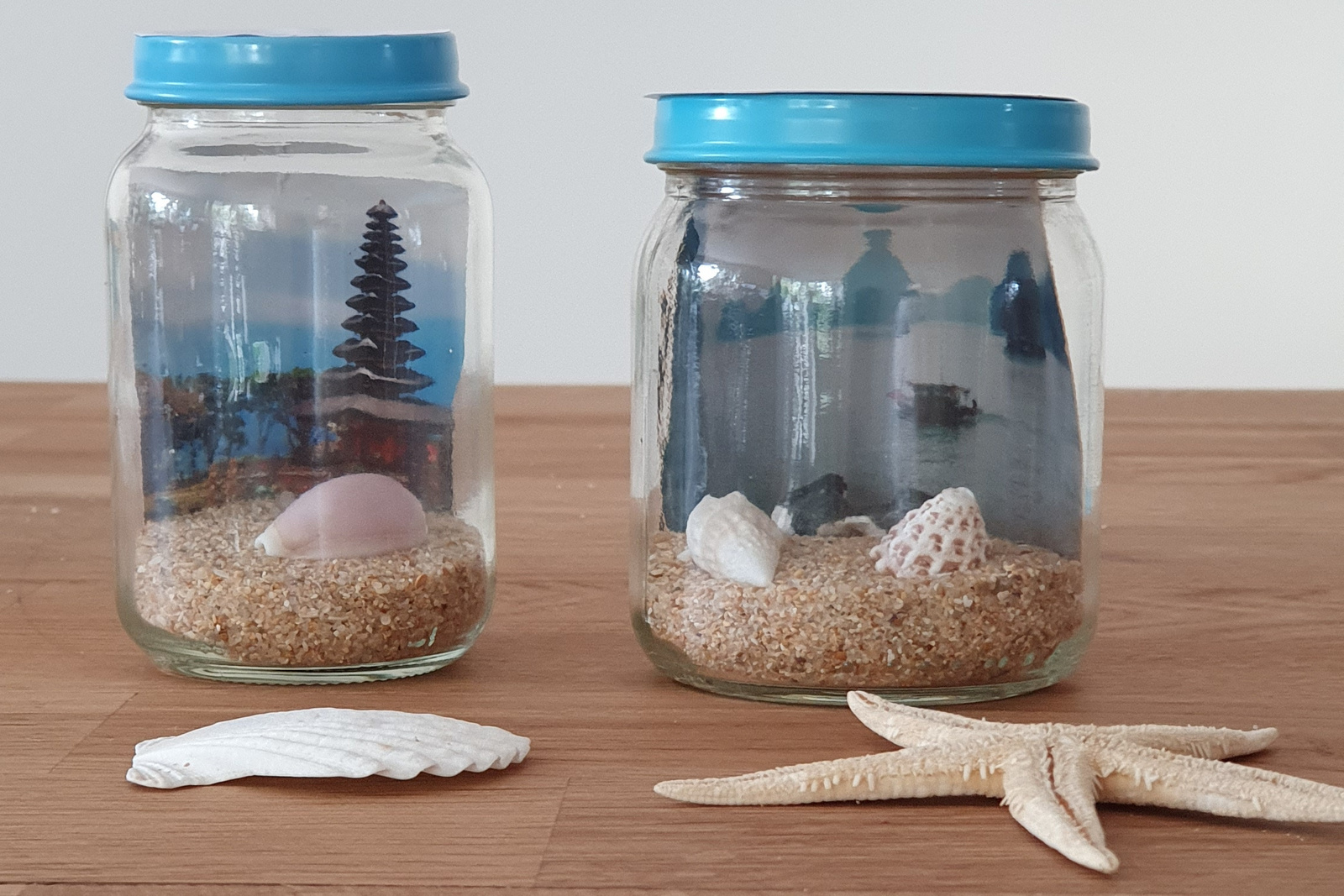 Zwei dekorative Gläser mit Muscheln und Sand, im Hintergrund eine stilisierte Landschaft, auf einem Holzuntergrund.