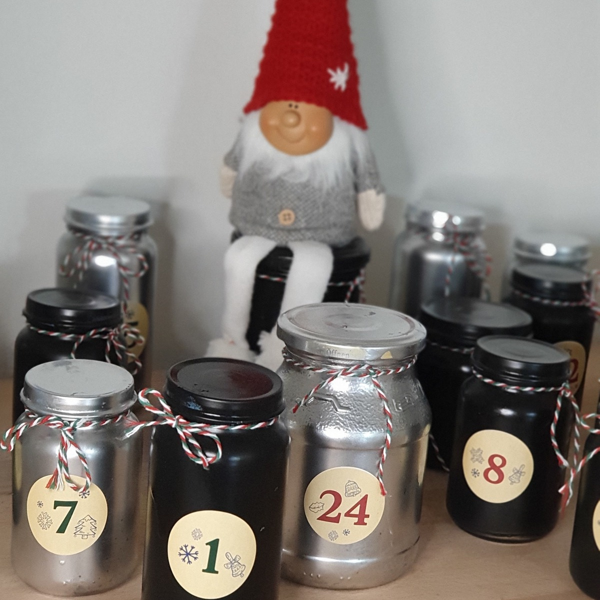 An einem Holztisch stehen mehrere Weihnachtsgläser in verschiedenen Farben, nummeriert von 1 bis 24, neben einer kleinen, sitzenden Weihnachtsfigur.