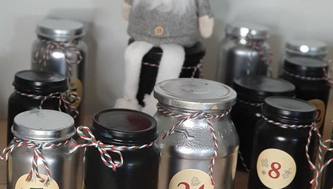 An einem Holztisch stehen mehrere Weihnachtsgläser in verschiedenen Farben, nummeriert von 1 bis 24, neben einer kleinen, sitzenden Weihnachtsfigur.