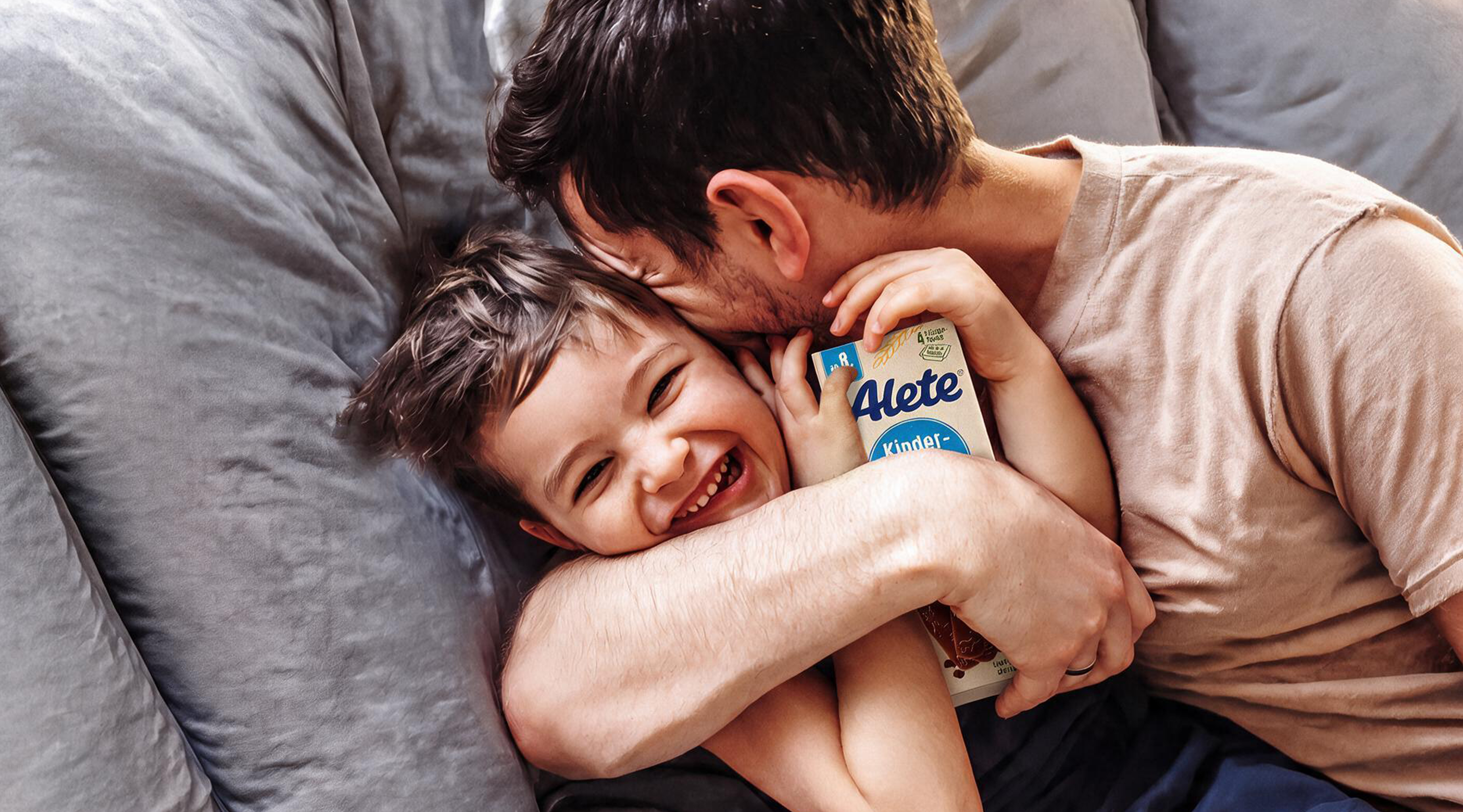 AI-generated: A father hugs his laughing son on a couch while the boy holds a package of Alete Kinderkeks in his hands.