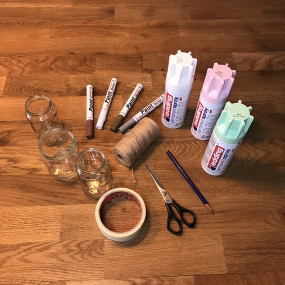 Bastelmaterialien auf einem Holztisch: Gläser, Marker, Sprühfarbe, Schnur, Klebeband und Schere.