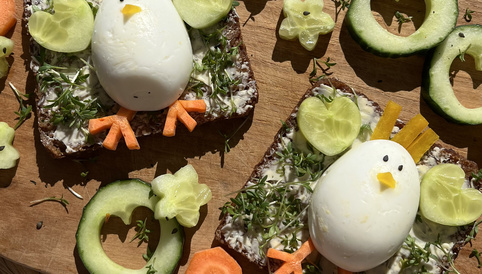 Dekoratives Sandwich mit einem gekochten Ei, das als Hühnchen gestaltet ist, umgeben von Gurken- und Karottenstücken sowie Kräutern auf einem Holzbrett.