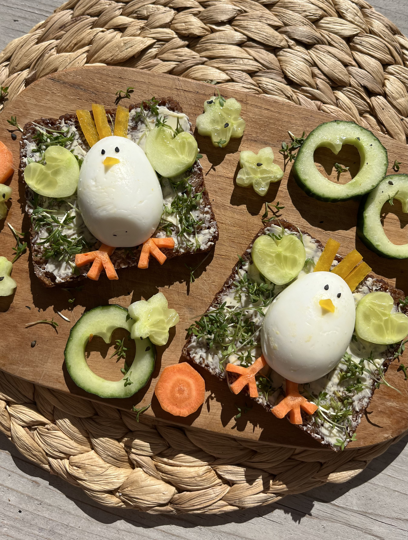Dekoratives Sandwich mit einem gekochten Ei, das als Hühnchen gestaltet ist, umgeben von Gurken- und Karottenstücken sowie Kräutern auf einem Holzbrett.
