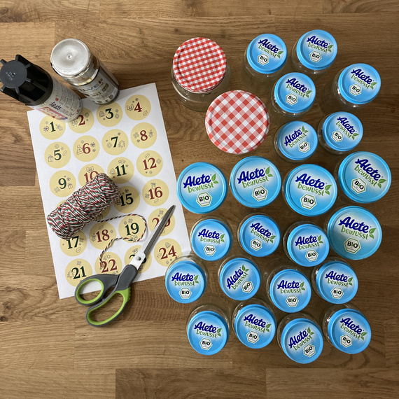 Bastelmaterialien für einen Adventskalender: Gläser mit bunten Deckeln, Aufkleber mit Nummern, Schere und Schnur auf Holztisch.