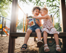 AI-generated Bild: Zwei fröhliche Kinder auf einem Spielplatz teilen eine Packung Alete Schnitties, während die Sonne durch die Bäume scheint.
