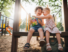 Zwei fröhliche Kinder auf einem Spielplatz teilen eine Packung Alete Schnitties, während die Sonne durch die Bäume scheint.