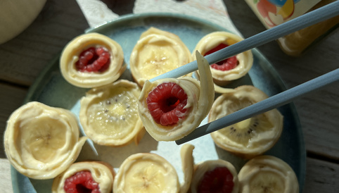 Bananen-Röllchen gefüllt mit Himbeeren auf einem grünen Teller, umgeben von frischen Himbeeren und einem Glas Alete Babybrei.