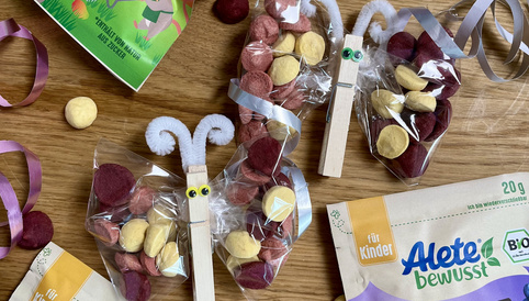 Bunte Alete Obsties für Kinder auf einem Holztisch, umgeben von Bastelmaterialien, wie kleinen Geschenken und bunten Bändern.