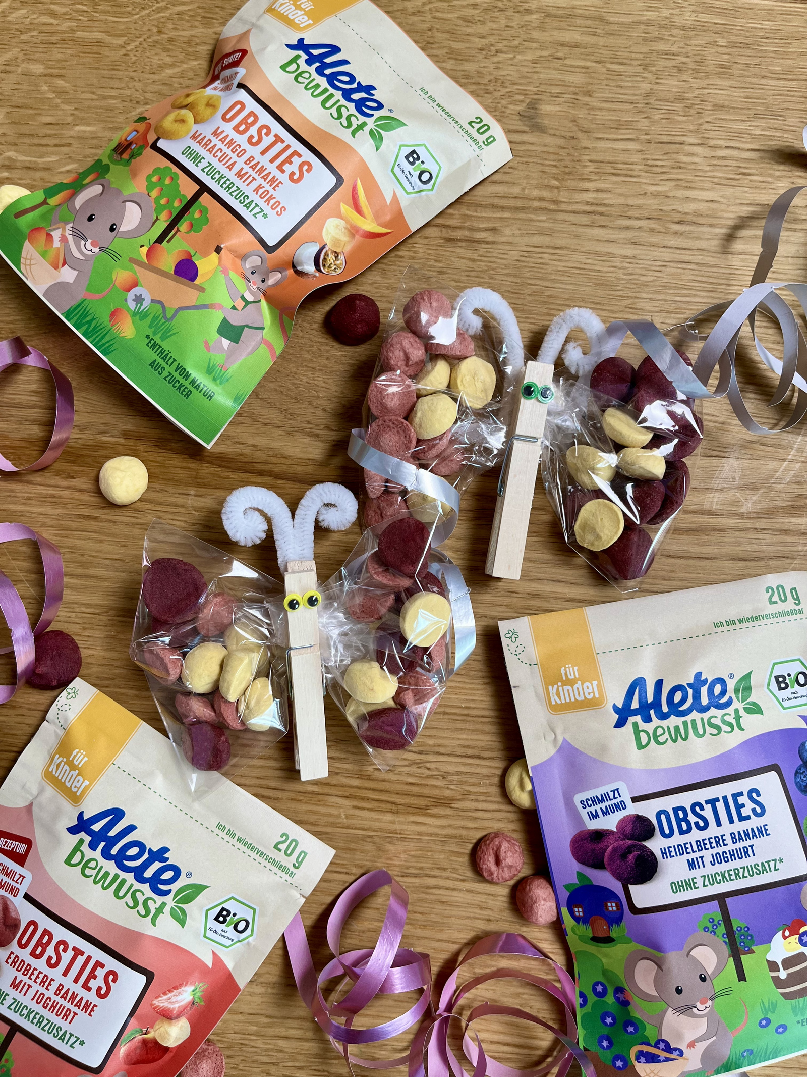 Bunte Alete Obsties für Kinder auf einem Holztisch, umgeben von Bastelmaterialien, wie kleinen Geschenken und bunten Bändern.