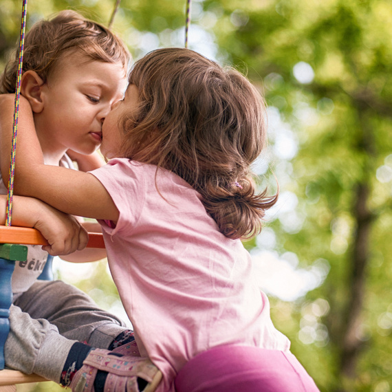 Zwei Kinder, ein Junge und ein Mädchen, küssen sich auf einer Schaukel im Freien, umgeben von Bäumen.