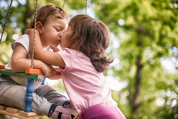 Zwei Kinder, ein Junge und ein Mädchen, küssen sich auf einer Schaukel im Freien, umgeben von Bäumen.
