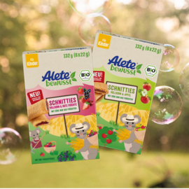 Bio-Snackpackungen von Alete "Schnitties" für Kinder, mit Sorten Vollkorn & Apfel sowie Vollkorn & rote Früchte, vor einer Naturkulisse.