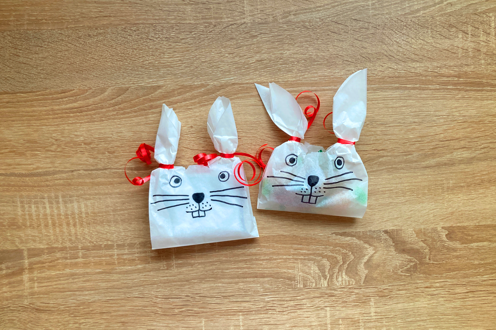 Zwei dekorierte Geschenktüten in Hasenform mit roten Schleifen und aufgemalten Gesichtern auf einem Holzuntergrund.