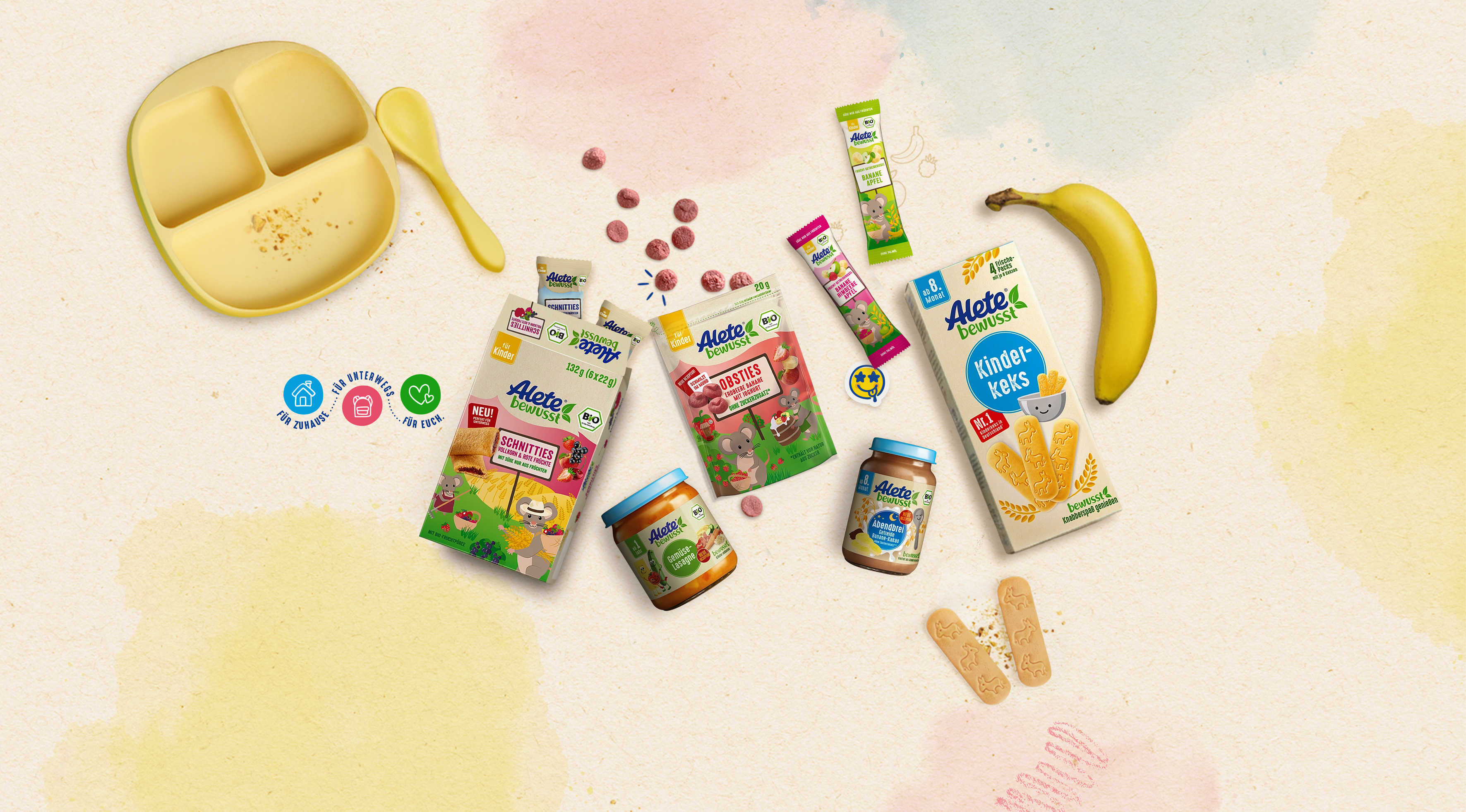 Bunte Auswahl an Baby- und Kleinkindernahrung von Alete: schnittige Snacks, Obst-Brei und Kekse, kombiniert mit einer Banane und einem Löffel.