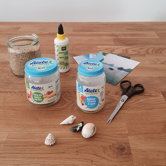 Zwei Gläser mit ALETE-Produkten und verschiedene Muscheln auf einem Holztisch, umgeben von Bastelmaterialien und Fotos.