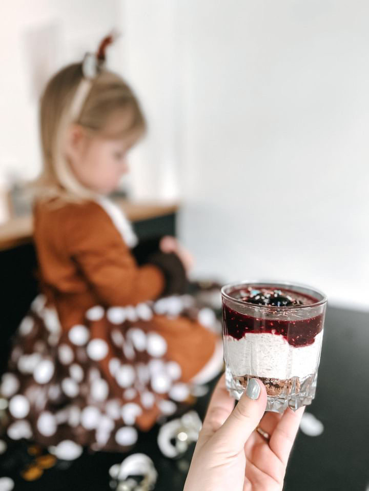 Hand hält ein Glas mit Schichtdessert in die Kamera - im Hintergrund sitzt unscharf ein Mädchen