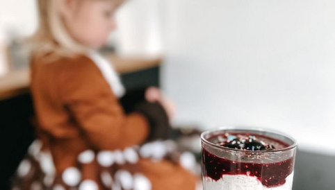 Hand hält ein Glas mit Schichtdessert in die Kamera - im Hintergrund sitzt unscharf ein Mädchen