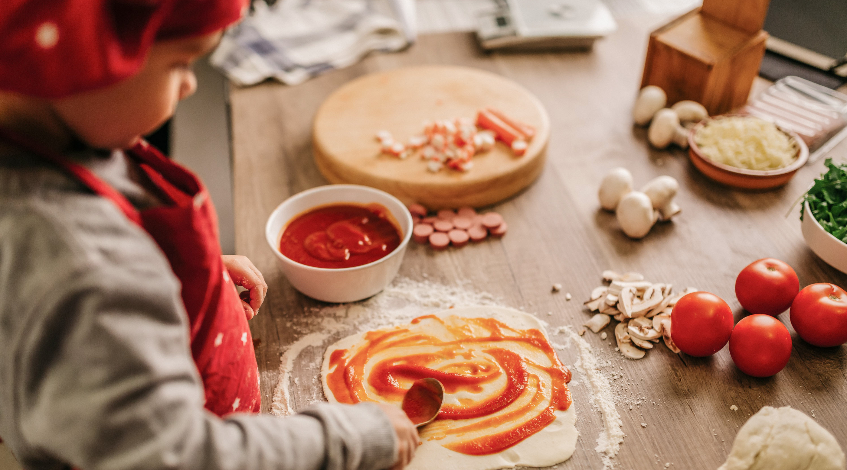 Kind in rotem Kochoutfit verteilt Tomatensauce auf einem Pizzateig auf einem Holzbrett, umgeben von frischen Zutaten.