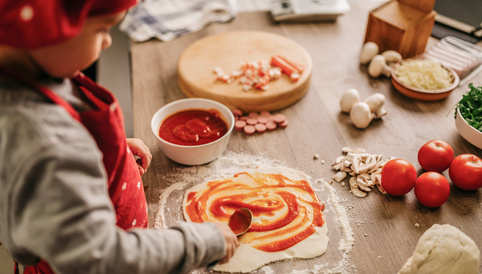 Kind in rotem Kochoutfit verteilt Tomatensauce auf einem Pizzateig auf einem Holzbrett, umgeben von frischen Zutaten.
