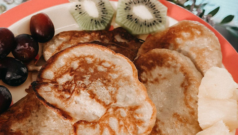 Goldene Pancakes auf einem Teller, umgeben von frischen Kiwi-Scheiben, Trauben und Ananasstücken, mit floraler Dekoration im Hintergrund.