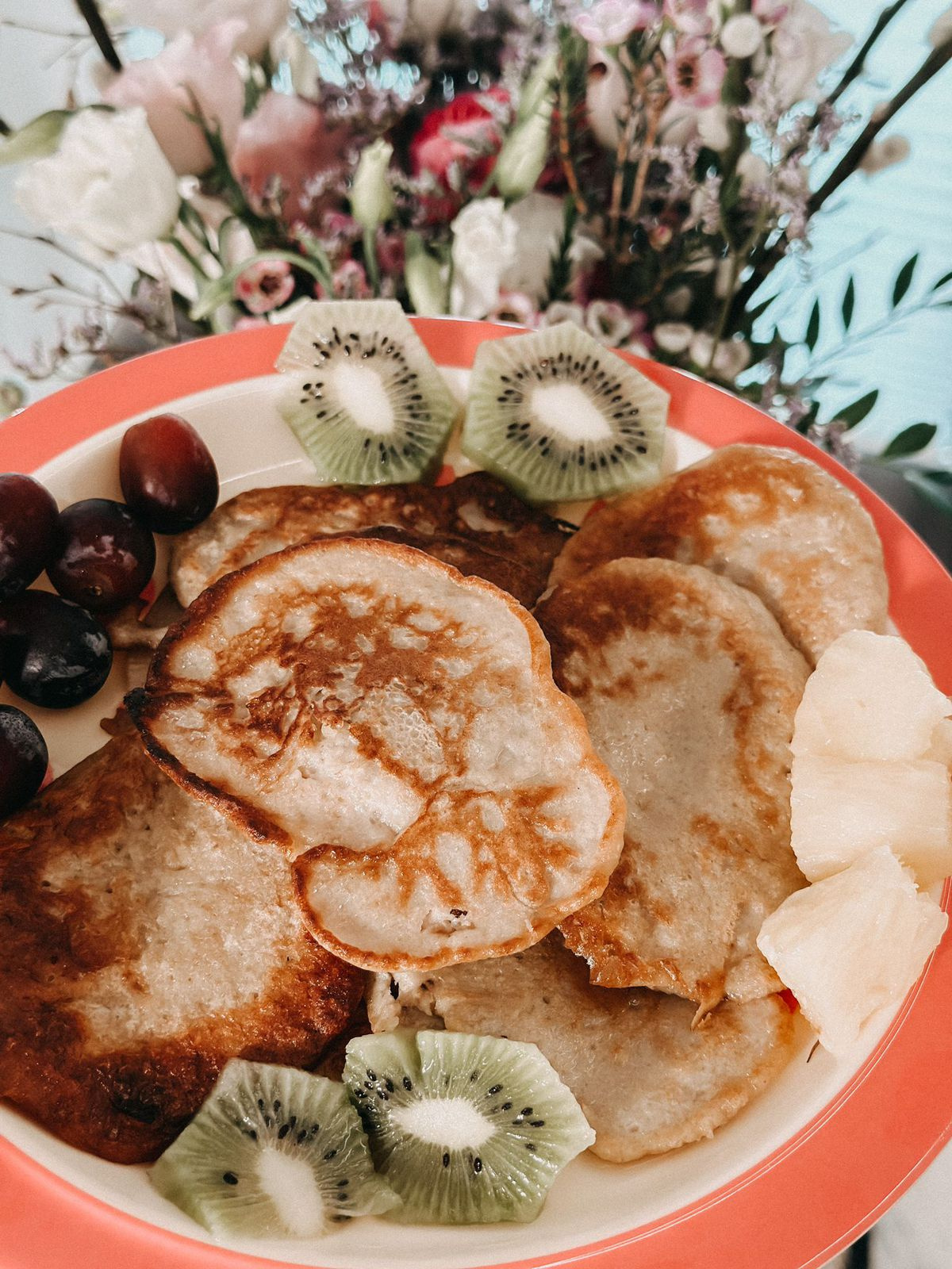 Goldene Pancakes auf einem Teller, umgeben von frischen Kiwi-Scheiben, Trauben und Ananasstücken, mit floraler Dekoration im Hintergrund.