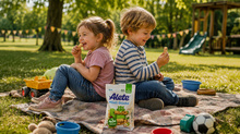 Zwei Kinder sitzen auf einer Decke im Park und genießen Alete Müsli Sticks. Im Hintergrund sind Spielzeug und eine Rutsche sichtbar.