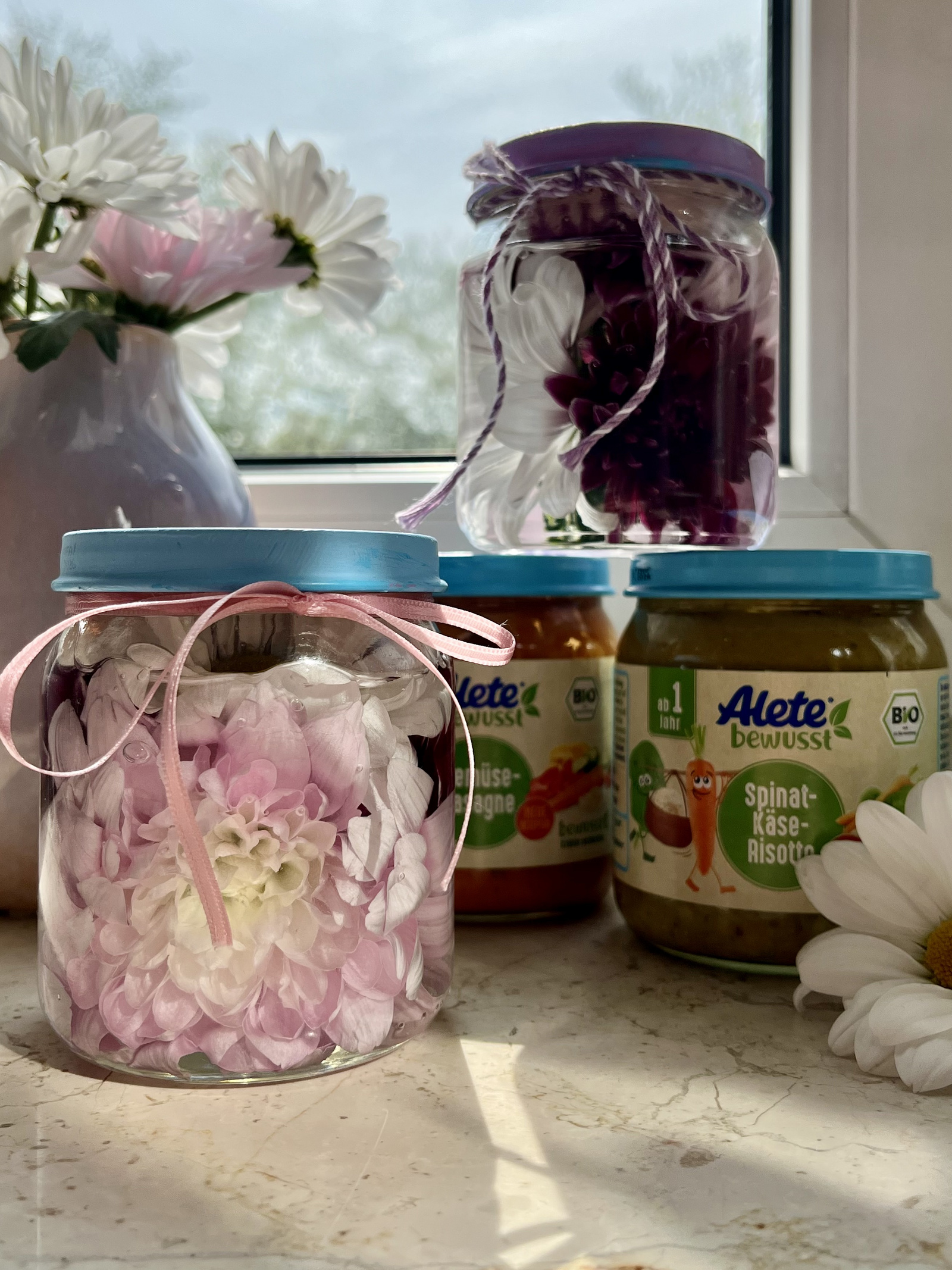 Gläser mit bunten Blumenarrangements und Babygläschen von Alete auf einer Fensterbank, umgeben von weißen Blumen.
