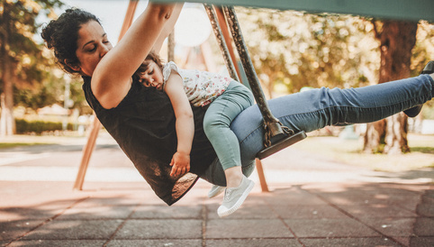 Mutter schwingt mit schlafendem Kind auf einer Schaukel im Park, umgeben von Bäumen und natürlichem Licht.