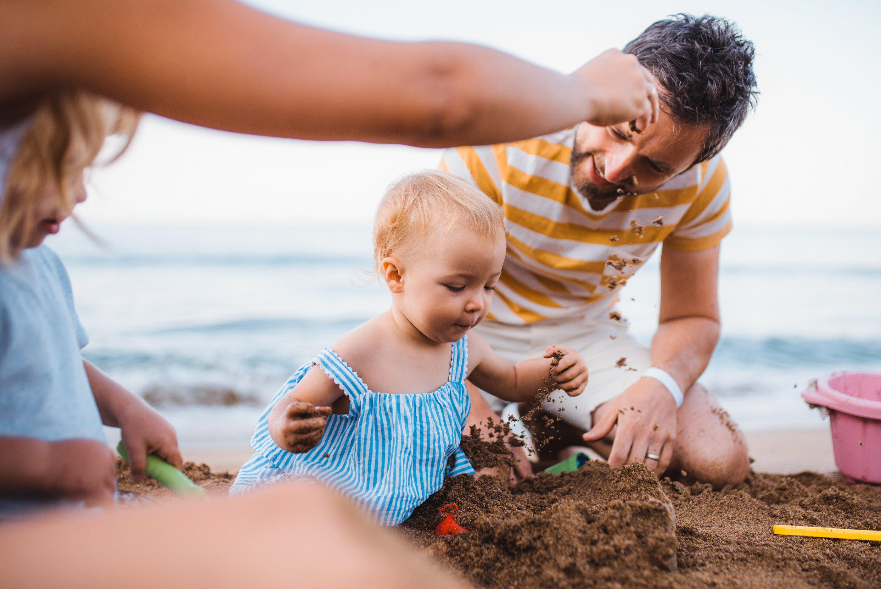Kind spielt glücklich im Sandstrand, während ein Erwachsener neben ihm mit Sand spielt. Meer im Hintergrund.