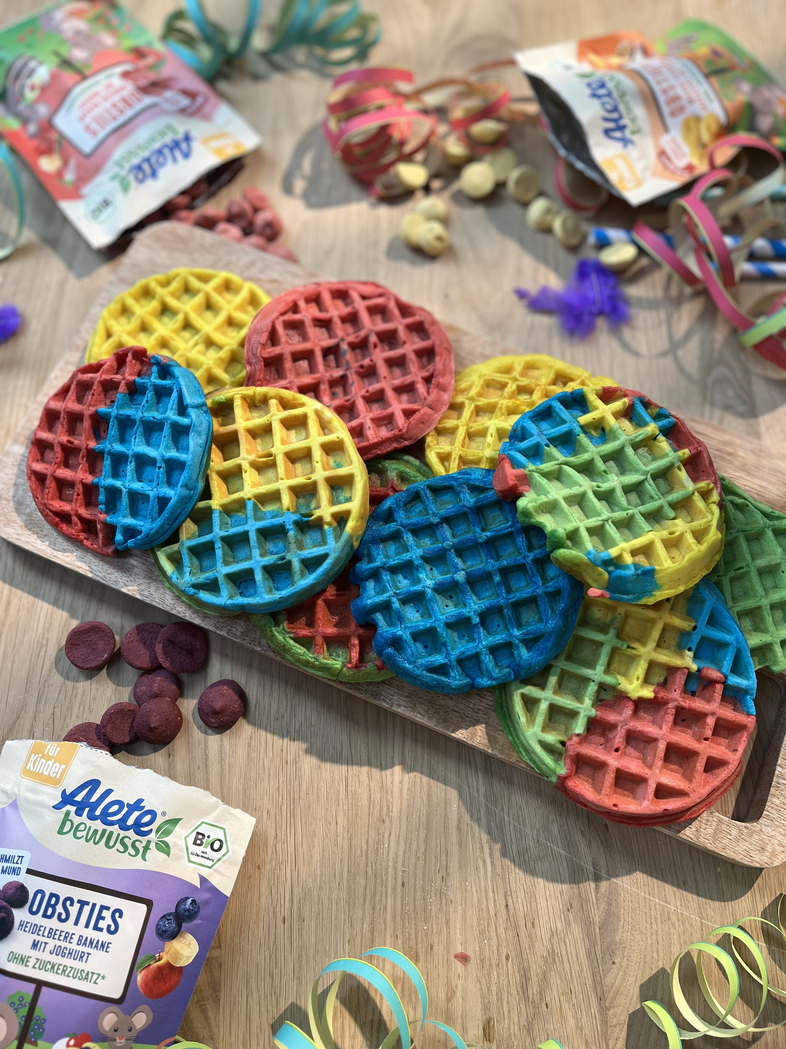 Bunte Waffeln auf einem Holzbrett, umgeben von Konfetti und Obstsnack-Verpackungen, ideal für Kindergeburtstage.