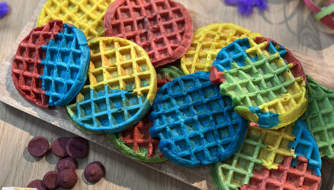 Bunte Waffeln auf einem Holzbrett, umgeben von Konfetti und Obstsnack-Verpackungen, ideal für Kindergeburtstage.