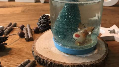 Weihnachtliche Schneekugel mit einem blauen Baum und einem Rentier. Dekoriert mit Zimtstangen und Holzstern auf einem Holzteller.