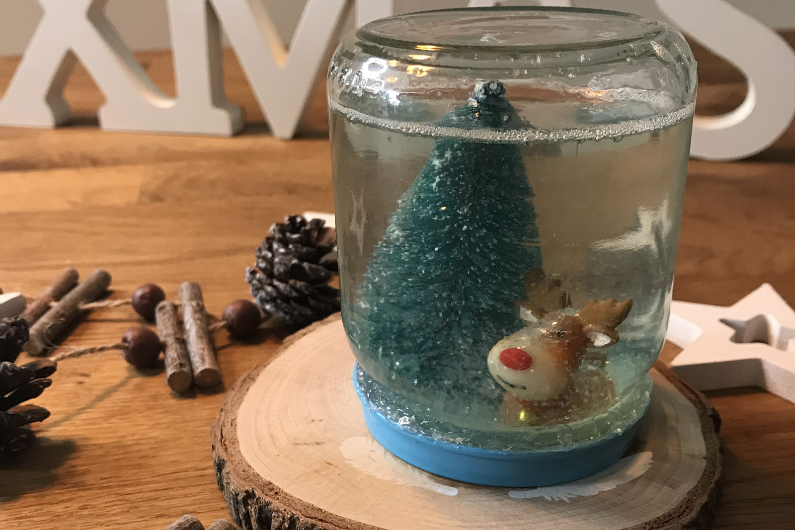 Weihnachtliche Schneekugel mit einem blauen Baum und einem Rentier. Dekoriert mit Zimtstangen und Holzstern auf einem Holzteller.