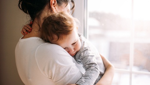 Eine Mutter umarmt ihr schlafendes Kind am Fenster, während sanftes Licht den Raum erhellt.