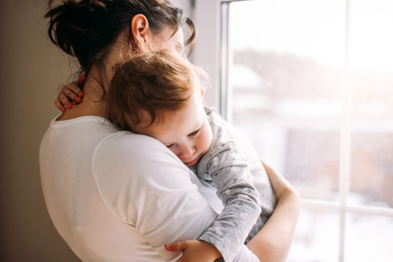Eine Mutter umarmt ihr schlafendes Kind am Fenster, während sanftes Licht den Raum erhellt.