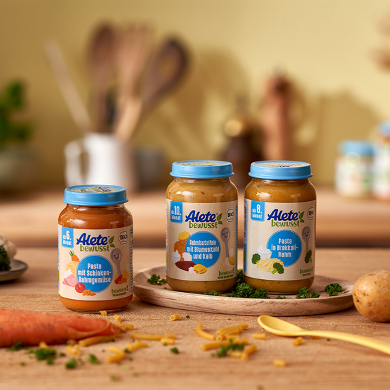 Drei Alete Gläschen auf einem Holztisch, eine Karotte und ein Löffel liegen davor