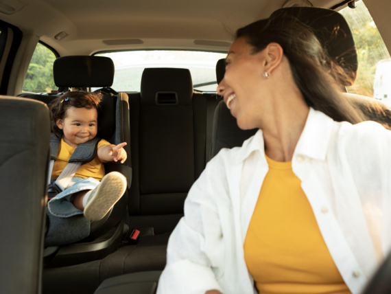 Lächelnde Mutter schaut auf ihr Kleinkind im Kindersitz während einer Autofahrt. Sonnenschein durch die Windschutzscheibe.