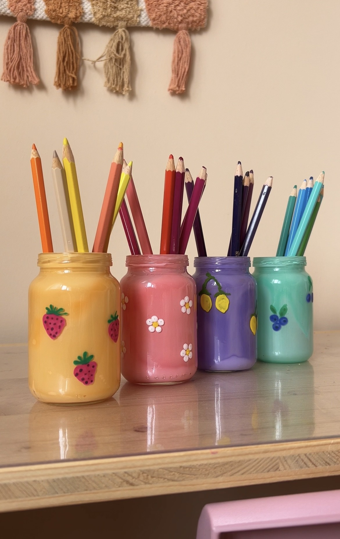 Bunte Gläser mit dekorativen Motiven, gefüllt mit Buntstiften in verschiedenen Farben, stehen auf einem Holzregal.