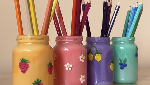 Bunte Gläser mit dekorativen Motiven, gefüllt mit Buntstiften in verschiedenen Farben, stehen auf einem Holzregal.