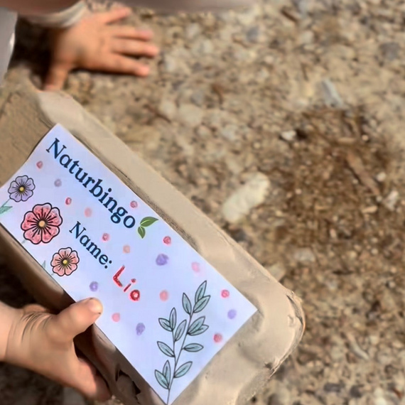 Kind hält ein Naturbingo-Kartenhalter mit dem Namen "Lio" und bunten Pflanzenmotiven in der Hand, auf einem schotterigen Untergrund.