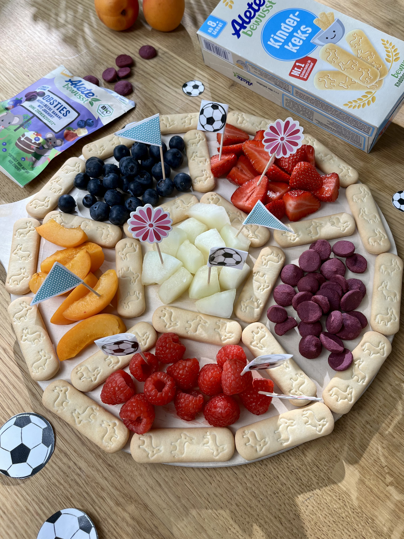 Bunte Obst- und Keksplatte mit Erdbeeren, Himbeeren, Blaubeeren, Melonen, Aprikosen und Keksen in Fußballform, dekoriert mit kleinen Fähnchen und Snacks.