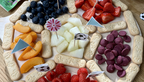 Bunte Obst- und Keksplatte mit Erdbeeren, Himbeeren, Blaubeeren, Melonen, Aprikosen und Keksen in Fußballform, dekoriert mit kleinen Fähnchen und Snacks.
