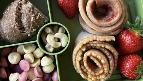 Gesunde Lunchbox mit Erdbeeren, Nussmix, bunten Obstsnacks und zwei gerollten Pfannkuchen mit Marmelade.