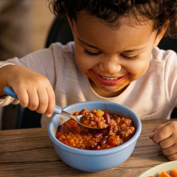 Ein lächelndes Kleinkind sitzt am Tisch und genießt eine Schüssel mit buntem Chili con Carne, während es mit einem Löffel darin rührt.