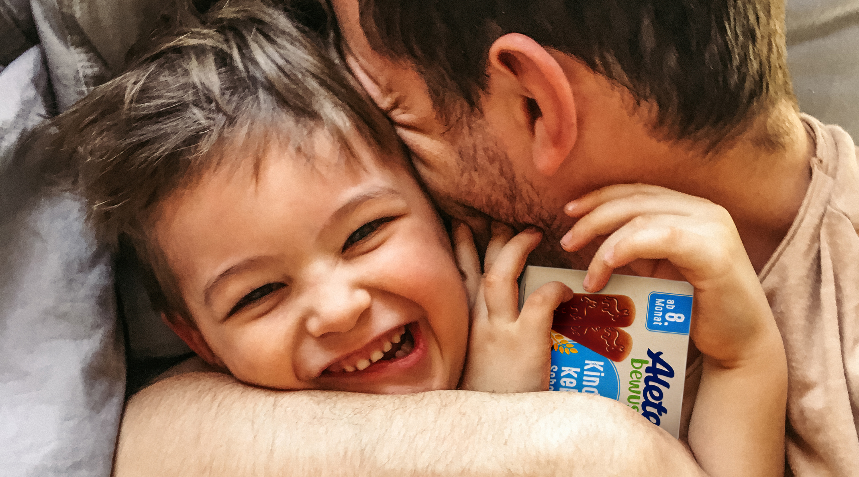 Vater umarmt lachenden Sohn im Bett, beide glücklich und entspannt, während der Junge eine Verpackung mit Alete Keksen hält.