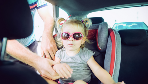 Ein kleines Mädchen in Sonnenbrille sitzt in einem Autositz, während eine Hand sie anschnallt.