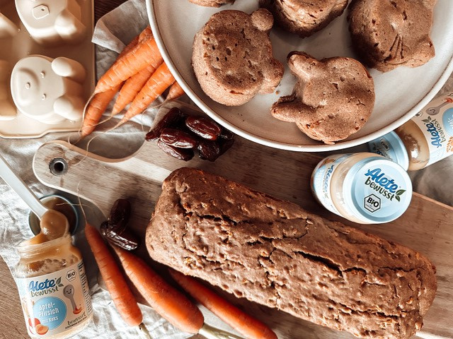 Karottenkuchen in Kasten- und Hasenform liegt auf Holzbrett, daneben Karotten und Alete-Gläschen